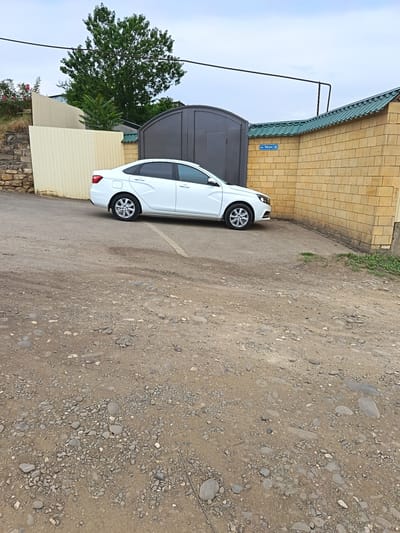 This media file depicts a white sedan parked on a gravelly, unpaved road in front of a wall. The wall is constructed of light-colored bricks and is topped with a green, wavy roof. To the left of the brick wall, there is a tall, dark-colored gate and a pale yellow fence. Behind the fence, a large green tree with dense foliage is visible. A small cluster of pink flowers is also present on the left side of the image, near a stone retaining wall.
A blue street sign with white text is affixed to the brick wall, reading "ул. Мира 10". The sky is overcast, suggesting it is daytime, possibly in the morning or late afternoon, with moderate weather conditions. The ground is covered in loose gravel and small stones, with some patches of dry earth. The scene appears to be a residential or semi-rural area in Kasumkent, Russia. There are no people visible in the image, and the focus is on the parked car and the surrounding environment. The overall impression is one of a quiet, everyday scene.
