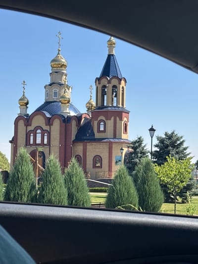 The image shows an Eastern Orthodox church with golden domes, a blue roof, and tan and red brick walls. The church is situated in a well-maintained outdoor setting with a clear blue sky, green grass, and several evergreen trees. A lamppost is visible to the right of the church. The foreground of the image is dominated by the interior of a vehicle, with a dark-colored dashboard or armrest in the lower portion. This suggests the photo was taken from inside a car looking out at the church. The church appears to be located in the village of Voronovo, Russia.