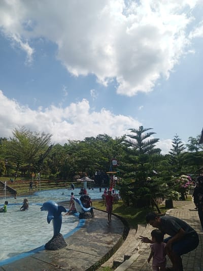 The image depicts a daytime scene at a public swimming pool with a water park area, likely located in Darsono, Indonesia, given the context. The sky is partly cloudy with bright sunlight. In the foreground, a blue dolphin statue stands on a dark rock base in the shallow end of the pool. To the right of the pool, a man is crouched down, interacting with a small child who is wearing a pink outfit and standing on a brick pathway. The pathway curves around the pool and is bordered by lush green grass and trees.
Further into the pool, several individuals, including children and adults, are seen swimming, wading, and playing. One child in a green shirt is visible on the left side of the pool. A larger blue dolphin statue is positioned in the water to the left of the man and child. In the background, more trees and vegetation surround the water park, with glimpses of structures and water features, suggesting a recreational park. The overall atmosphere appears to be one of leisure and family enjoyment on a sunny day.