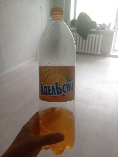 A hand holds a clear plastic bottle containing an orange beverage with a light brown cap. The bottle has a label with Russian text that reads "АПЕЛЬСИН" (Apelsin, meaning orange) and an image of an orange slice. The background shows a radiator and a window, suggesting an indoor setting in a city like Borodino, Russia. The lighting indicates it is daytime, possibly with sunlight coming through the window.