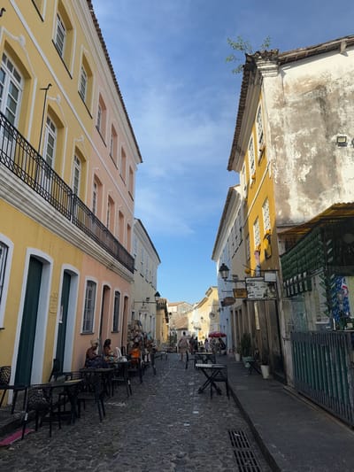 Cobblestone street between tall buildings. The buildings are painted in shades of yellow and pink, with white trim and window frames. Black metal balconies and ornate detailing adorn the façades. Tables and chairs line the street, occupied by people, suggesting a café or restaurant area. The sky is visible between the buildings, a pale blue with scattered clouds. The perspective is looking up the street, creating a sense of depth.