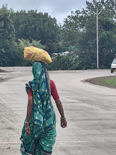 In Walsawangi, India, a person wearing a teal and green patterned sari with a red blouse walks on a paved road. They are carrying a large, yellow cloth bag balanced on their head. The road curves to the right, with a grassy embankment on the right side. In the background, a white vehicle is visible, and dense green trees line the horizon under a cloudy sky. A utility pole with wires is also present. The person is adorned with several bangles on their wrists.
