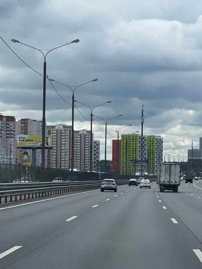 The image shows a multi-lane highway, likely an overpass, with traffic moving along it. The setting is in Novodrozhzhino, Russia, as indicated by the Russian text on a large advertising banner visible alongside the highway. The sky is overcast, with gray clouds dominating the upper portion of the picture.
Several tall apartment buildings are visible in the background, painted in different colors (red, green, white, etc.) and arranged in rows. Streetlights line the side of the highway. Cars, a truck, and a van are traveling on the highway.