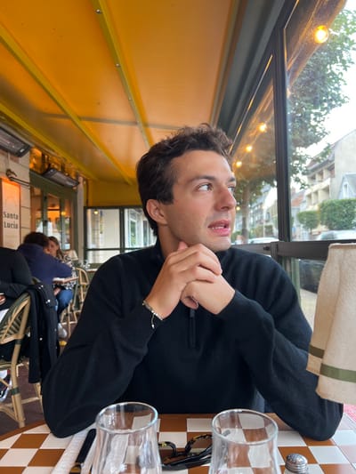 The image depicts a young adult male seated at an outdoor or semi-enclosed restaurant terrace, identified as Santa Lucia in Deauville, France, based on a visible sign. The man, with dark wavy hair and light stubble, wears a black quarter-zip sweater and a light-colored bracelet on his left wrist. He is positioned at a table with a brown and white checkered surface, looking towards the right of the frame with his mouth slightly open, his hands clasped in front of him.
On the table in the foreground are two clear drinking glasses, a pair of tortoiseshell-patterned eyeglasses, a silver salt shaker, and a folded white napkin with a green stripe. A dark-handled utensil is also present.
The background to the left reveals a yellow awning covering the terrace and two dark-colored infrared heaters mounted overhead. Other patrons are visible further back, seated at tables with wicker chairs. A white sign with "Santa Lucia" in red script is partially visible on a wall. To the right, beyond a large glass window, is an outdoor street scene with buildings, trees, and a cloudy sky. Small, warm-toned light bulbs are strung along the window's upper frame.