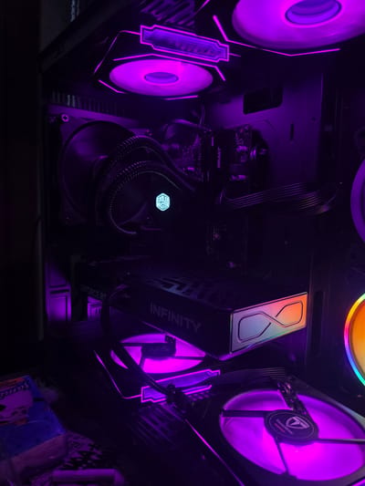 The image shows the interior of a computer tower, illuminated by purple LED lights. Components visible include fans, a graphics card labeled "GEFORCE RTX INFINITY," a CPU cooler with a Cooler Master logo, and RAM sticks labeled "DDR4 BEAST FURY." There is also a small, blue and white package with a cartoon image on the lower left. The scene is dark, with the bright purple lights highlighting the computer hardware. The location of Aleksandrovo, Bulgaria is not depicted or relevant to the image content.
