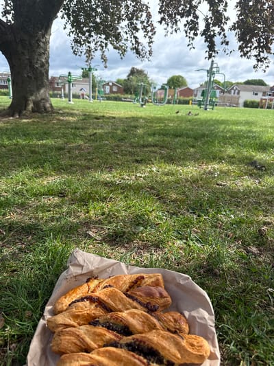 A photograph taken at ground level in High Wycombe, United Kingdom, captures a scene within a park. In the foreground, a brown paper bag containing a pastry, possibly a braided chocolate danish, sits on the grass. The pastry is golden-brown with visible layers and a dark filling. The grass is green and interspersed with patches of bare earth and dry leaves. In the midground, the grass stretches towards a row of houses in the background. There is outdoor exercise equipment visible, including green metal structures, suggesting a public park or recreation area. A tree with dark, leafy foliage is visible to the left, partially framing the scene. The sky is overcast with grey clouds. There are no people visible in the image. The scene suggests a peaceful outdoor setting, possibly during a picnic or break.