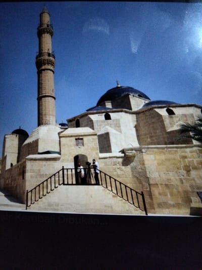 A tall, slender minaret made of tan stone stands to the left of a mosque with a dark blue dome. The mosque is constructed of light-colored stone and features arched windows. A set of stairs with a black railing leads up to an entrance where two individuals are visible. One person, wearing a red garment, stands near the stairs, while another person in a white garment is positioned further back, holding a camera or video equipment. The sky above is a clear, bright blue. The lower portion of the image shows a dark band with faint white text, possibly a watermark or caption, reading "si Kembali Dibukan...". The scene is likely outdoors, given the clear sky and the presence of a palm frond visible on the right side of the mosque.