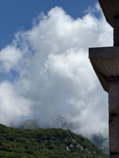 The image captures a breathtaking view of a mountainous landscape in Kubasi, Montenegro, seen from a high vantage point. The top of the frame is partially obscured by a dark, textured wooden structure, possibly a beam or a roof edge, suggesting the view is from a building or balcony.
The scene is dominated by a dramatic interplay of natural elements. On the right, a steep, densely forested mountainside descends vertically, covered in lush green trees and rocky outcrops. To its left, a deep valley is engulfed in swirling white and grey mist, which partially conceals the lower parts of distant, rugged mountain peaks. Through the ethereal haze, glimpses of stark, grey rocky cliffs and slopes are visible, hinting at the scale and complexity of the terrain beyond.
The lower-left portion of the image reveals a clear blue sky, interspersed with fluffy, bright white cumulus clouds, indicating fair weather at higher elevations. The contrast between the clear sky and the misty valley suggests dynamic weather conditions typical of mountainous regions.
The time of day appears to be bright daytime, with ample natural light illuminating the clouds and the visible parts of the mountains. There are no visible people, animals, or man-made objects other than the wooden structure in the immediate foreground. No text is visible in the image. The overall impression is one of majestic natural beauty and the vastness of the mountain environment.