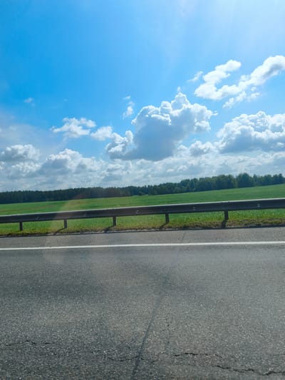 The image captures a scene from the side of a road in the Драчкаўскі сельскі Савет, Belarus. The foreground is dominated by the asphalt road surface, marked with a white line indicating a lane division. A metal guardrail runs along the edge of the road, separating it from a grassy area. Beyond the guardrail, a vibrant green field stretches towards a dense line of trees forming the horizon. The sky above is a brilliant blue, dotted with fluffy white cumulus clouds, suggesting a clear and pleasant day. The overall impression is one of a rural roadside landscape under a bright sky. There are no people or vehicles visible in the frame.