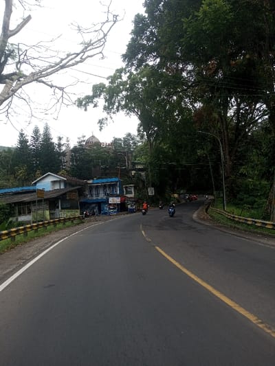 The image displays a two-lane asphalt road curving to the right within a hilly, heavily vegetated area, identified as the city of Sat Thay (East), Myanmar. The sky is uniformly overcast.
On the left side of the foreground, a yellow and black striped guardrail runs parallel to the road, which features a solid white line on its left edge and a continuous yellow center line, indicating a no-passing zone. Beyond the guardrail, a series of multi-story buildings, some with prominent blue and white facades, are constructed into the sloping terrain. Foreign language text is visible on various signs attached to these structures, with some legible words appearing to include "JUALAN SEJAK 2023" and "MINUMAN". Above these buildings and partially obscured by evergreen trees, a larger, more elaborate building features a distinct dome-shaped roof and additional text, possibly "HOTEL" or "IDEAL", on its facade.
Several individuals are operating motorcycles on the road, all traveling away from the viewer along the curve. A red motorcycle is positioned near the center line, followed by a dark-colored motorcycle, and two additional red motorcycles further down the road. A red passenger vehicle is partially visible behind the last motorcycle.
The right side of the road is densely bordered by tall green trees and abundant foliage. A street light pole and overhead utility wires are visible among the tree canopy. Another section of yellow and black striped guardrail is present on the right shoulder further down the curve.