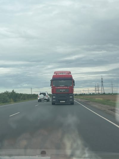 An exterior, eye-level, full shot shows a red semi-truck on a highway in Zorinskii, Russia. The truck is in the foreground and is facing forward. The truck has "MAN" written on the front and a license plate that reads "r003ee 64". There are two white vehicles on the road ahead of the truck, a smaller SUV and a white passenger car. The road is asphalt with a white line marking the lanes. The environment is rural, with fields and trees on either side of the road and power lines in the distance. The sky is cloudy and overcast.