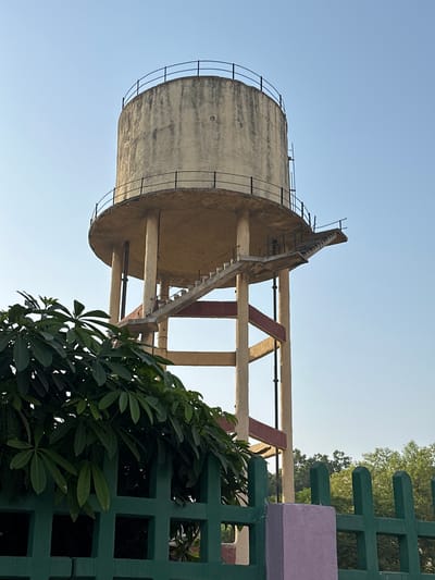 The image features a concrete water tower against a clear blue sky. The tower is cylindrical with a ladder leading to the top. It is supported by concrete beams, some reinforced with red paint. To the right of the tower, a green fence with square cutouts is partially obscured by leafy plants. The location is likely Mathura, India. There are no people visible.