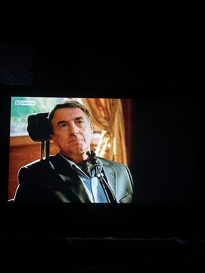 The image shows a close-up of a man wearing a suit and a blue shirt. He is seated in a chair with a headrest. He has a concerned expression on his face. A logo that reads "b cinema" is visible in the top left corner of the image. The background appears to be a room with curtains. The image is of a broadcast or recording, not a scene from the city of Kashin, Bulgaria.
