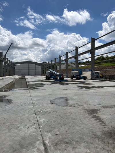 An outdoor construction site in Janowice, Poland is depicted. The scene shows a partially completed building structure with exposed steel beams. In the foreground, a wet concrete surface is visible with several puddles. A blue articulated boom lift, identified as a "Genie Z-60/60," is positioned in the middle ground. Two workers wearing orange vests are operating from the platforms of two different boom lifts. One boom lift is to the left and the other is to the right of the central boom lift. A large, unfinished building with a white facade stands in the background. The sky is bright blue with scattered white clouds. A construction crane arm extends from the left side of the image into the upper sky. The surrounding landscape includes a vegetated hill to the right.