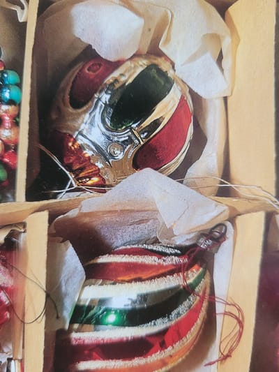 The image displays a close-up view of Christmas ornaments nestled in their packaging. The top ornament is a round, multi-faceted bulb with sections of red, green, and silver. It is partially wrapped in off-white tissue paper. Below it, another ornament, also round, features alternating stripes of red, white, and green, with a reflective surface. This ornament is also situated in protective packaging, with some red string visible. The background consists of cardboard dividers and the muted tones of the packaging material. The image provides no contextual information about the city of Gävle, Sweden, or any people or actions within the scene.