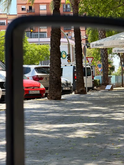 The image, taken in Castelló De La Plana, Spain, captures a street scene through a rearview mirror, as suggested by the dark frame. In the foreground, a grey tiled sidewalk is visible, dappled with sunlight and scattered with green leaves. Several cars are parked on the street, including a red Ford Focus with license plate 8945 CZJ, a silver BMW X3, and a dark grey Volkswagen Golf with license plate 4301 NMG. Beyond the cars, a white van is parked.
The background features palm trees, a building with multiple stories and balconies, and awnings displaying the "Alhambra" logo, indicative of a bar or restaurant. A traffic light with a yellow signal is also visible. The overall impression is a sunny day in an urban environment, likely a commercial or residential street with parking available.