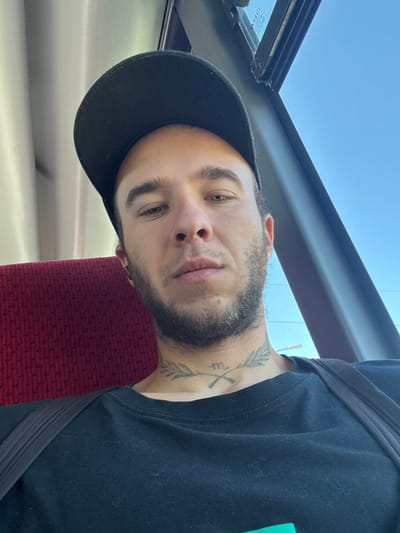 A close-up, selfie-style shot shows a light-skinned male from the chest up, wearing a black t-shirt and a black baseball cap. He is seated, with a dark gray strap visible across his chest. A textured, crimson red seat is to his left. His face is partially obscured by the cap, but his eyes, nose, and mouth are visible. He has a short beard and a tattoo on his neck that appears to be a laurel wreath with a symbol in the center. To the right of the man, a window reveals a clear blue sky and a dark gray frame. The overall lighting suggests it is daytime. The setting appears to be inside a vehicle, possibly a bus or train, given the seat and window. The background suggests an outdoor environment consistent with the city of Šestajovice, Czechia, under a clear sky.