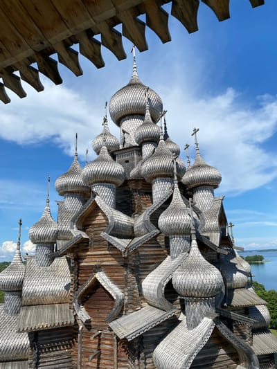 The image shows the wooden Church of the Transfiguration on Kizhi Island in Boyarshchina, Russia. The church is a prominent example of traditional Russian wooden architecture, characterized by its numerous onion domes, intricate woodwork, and multi-tiered design. The sky is blue with scattered white clouds. In the background, a body of water and green trees are visible, suggesting a natural or park-like setting. The foreground shows the underside of a wooden structure, possibly a balcony or roof overhang, with carved wooden elements. The image is taken from a low angle, looking up at the church's complex roofline and domes.