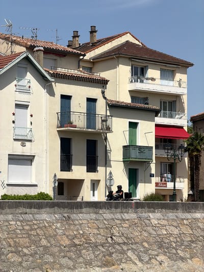 This image captures a street scene in Mercurol-Veaunes, France, on a clear, sunny day. In the foreground, a sturdy stone wall forms a retaining structure, with its rough-hewn stones laid in a diagonal pattern and secured by concrete mortar. A low green hedge runs along the top of this wall, bordering a paved area.

Dominating the midground are several multi-story buildings with pale yellow facades and red tile roofs. The architecture features balconies with black metal railings, several with doors opening onto them. Some windows are covered with white rolling shutters. On the left, a white building with a green-trimmed roof and a smaller balcony adorned with decorative elements is visible. In front of one of the buildings, a person wearing a helmet is seated on a scooter, with two black bags behind them. A street lamp with two lanterns stands near the right side of the buildings. A red awning extends over a balcony on one of the buildings. The background consists of a clear blue sky.