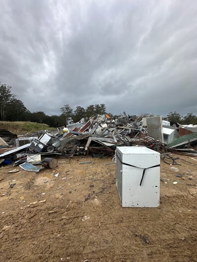 The image depicts a large pile of scrap metal and discarded appliances in an outdoor junkyard. The foreground is dominated by muddy ground with tire tracks, suggesting recent vehicle activity. A white appliance, possibly a washing machine or dryer, stands prominently in the mid-ground, secured with black straps. Behind it, a vast heap of mangled metal, corrugated iron sheets, and various machinery parts forms a chaotic landscape stretching towards a tree line under a cloudy, overcast sky. The scene is devoid of people and any discernible signs of urban infrastructure, placing it in a rural or industrial outskirts setting. The specific location in Raleigh, Australia, is not visually indicated by any landmarks or signage within the frame.