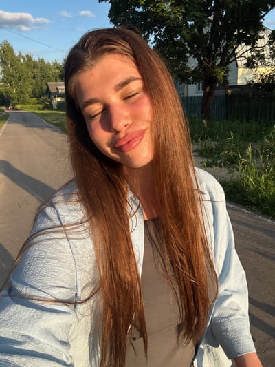 A young woman with long, auburn hair is captured in a selfie taken outdoors. She is wearing a light blue, textured jacket over a grey top. Her eyes are closed, and her head is tilted back, with her lips slightly parted in a relaxed expression. The sunlight casts a warm glow on her face, suggesting it is either early morning or late afternoon.

The background indicates a rural or suburban setting. To the left, a large tree with green foliage dominates the scene, with part of a building visible behind it. The building appears to have a corrugated metal exterior. Further in the background, more trees and greenery are visible, and a paved road or path curves through the landscape. The sky is a clear blue with a few white clouds. The overall impression is one of a casual, outdoor moment, possibly during a walk or a break in a sunny day. The location context is Шевлягино, Russia. There is no visible text in the image.