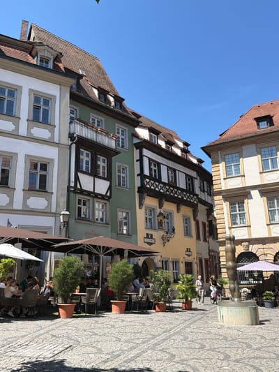 The image depicts a sunny day in the city of Bamberg, Germany. In the foreground, a cobblestone plaza is sparsely populated. To the left, outdoor seating areas of restaurants are visible, with a few patrons seated at tables beneath large umbrellas. Two tall potted trees stand prominently in the foreground, adding greenery to the scene.
The background features a row of historic European buildings with pitched tile roofs. The architecture is varied, with some buildings exhibiting timber-framing and others featuring classical facades. One building, painted a pale yellow, displays signage for "BRASSERIE." Another, painted green, has a balcony with a white railing. On the right side of the image, a modern art sculpture, resembling a stylized human leg, stands on a pedestal in the plaza.
In the middle ground, several people are engaged in various activities. Some are seated at the restaurant tables, while others walk across the plaza. A woman is seen riding a bicycle. The overall impression is one of a lively, picturesque urban square bathed in bright sunlight.