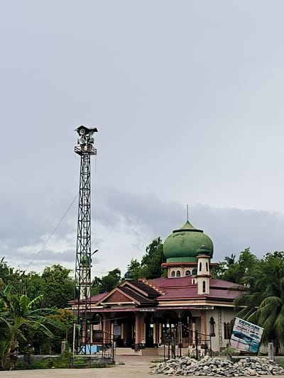 This vertical shot captures a mosque and a tall loudspeaker tower under an overcast sky, likely in Mesjid Gogo, Indonesia, as indicated by the location context.
The prominent subject is a mosque, positioned in the mid-ground, featuring a large, weathered green dome and a smaller matching dome. A slender, cream-colored minaret with a small domed top stands adjacent to the main structure. The mosque building itself has a reddish-brown tiled roof and cream-colored walls, with an arched entrance and architectural details suggesting a traditional Islamic design. Lush green tropical foliage, including palm trees and banana plants, surrounds and partially frames the mosque.
To the left of the mosque stands a tall, dark metal lattice tower. Perched at its apex is a platform supporting numerous horn-shaped loudspeakers, some sheltered by a small roof-like structure. A wire or cable runs diagonally from the tower towards the lower left.
The foreground consists of a relatively clear, unpaved or dirt area, with a substantial pile of light-colored rocks to the right. On the far right, near the rocks, a tilted white sign with blue text is visible. The most legible word on the sign is "DONASI," implying a request for donations. Below "DONASI," there are smaller, illegible words and what appears to be an image of vehicles. The sky is uniformly light grey and cloudy, suggesting a daytime scene without direct sunlight. There are no people visible, and no specific activities are taking place.