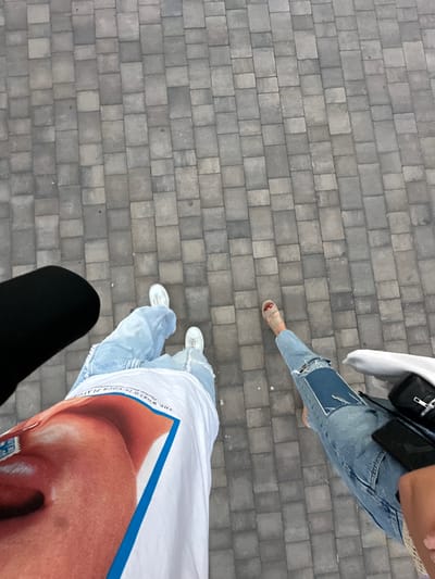 The image shows a ground-level view of a paved surface, likely a sidewalk, with two individuals and a portion of a third person visible. The pavement consists of gray, rectangular blocks arranged in a grid pattern.
The person in the upper right wears light blue denim jeans with a patchwork design, paired with tan-colored sandals. They carry a white bag and a black purse.
The second person wears light blue jeans and white sneakers and a white t-shirt with a face printed on it.
In the lower left corner, a person wearing black leggings appears, implying their presence.
The image was taken in Santa Pola, Spain. The perspective of the photo is from directly above.