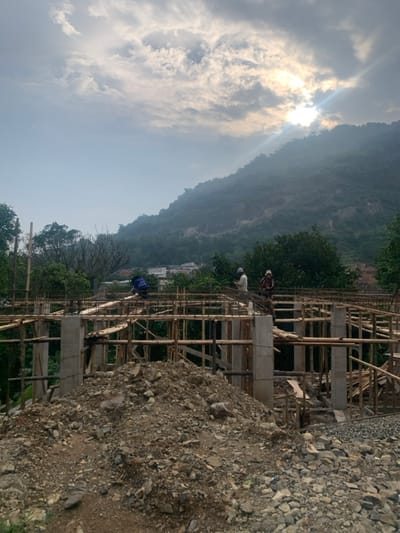 The image shows a partially constructed building in the city of Lagadar, Indonesia, at coordinates -6.927 latitude and 107.536 longitude. Three men are working on the construction site, two of whom are standing and one is bending over, seemingly working on the framework. The building is made of wood and concrete, with scaffolding surrounding it. The surrounding environment consists of a hillside covered with vegetation and a pile of loose gravel and dirt in the foreground. The sky is overcast with clouds, and the sun is shining brightly behind the clouds, creating a hazy effect.
