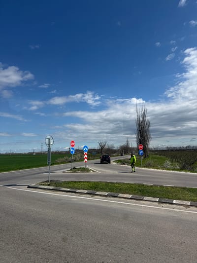 The image shows a rural road in Sitovo, Bulgaria. The sky is predominantly blue with scattered white clouds. A two-lane road stretches into the distance. A worker wearing a reflective yellow vest is standing on the left side of the road. A dark-colored car is also present on the road, heading towards the viewer. Several road signs are posted along the roadside, including a stop sign, turn signs, and signs with arrows. The road is flanked by green grass.