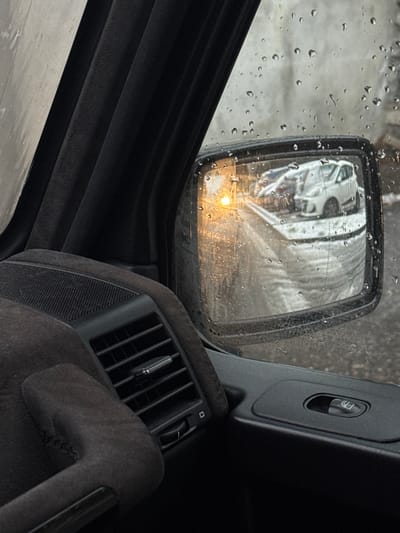 The image depicts a scene from inside a vehicle located in Lipomo, Italy, looking forward and towards the passenger side on a rainy day. Numerous water droplets adhere to the exterior of the window and the side mirror. Inside the vehicle, a dark fabric A-pillar is visible on the left, along with a dark, textured dashboard section featuring a rectangular air vent with horizontal slats and a control dial below it. A dark plastic door panel includes a window control switch. The external side mirror reflects a light-colored, possibly white, hatchback parked ahead, with another vehicle partially visible behind it. An amber-colored light, likely a turn signal or hazard indicator from the viewing vehicle, is prominently reflected in the mirror. A low, light-colored barrier or curb is also reflected in the lower portion of the mirror, separating the road from the parked vehicles. The background visible through the wet window and faintly in the mirror appears to be a light-colored, possibly textured, wall or building.