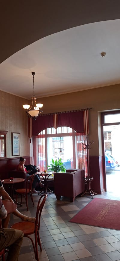 The image depicts the interior of a cafe, with a prominent arched ceiling and a large window at the back. A woman is seated at a table near the window, looking at her phone. She is wearing a red top and dark trousers. In front of her is a small round table with a glass of wine and a plate. The cafe has red upholstered seating, a coat rack, and tiled flooring. Outside the window, a blue car is visible parked on the street, with buildings and a doorway in the background. The overall impression is of a cozy and well-lit cafe. There is no indication within the image itself that it is located in Viljandi, Estonia.