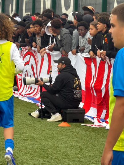 The image depicts an outdoor sporting event in Käerjeng, Luxembourg, from a low perspective on an artificial turf field. In the center foreground, a male photographer with a beard, wearing a black baseball cap backward, a black hoodie with a lion graphic on the back, black pants, and white Puma sneakers, is squatting or sitting on a black plastic case. He is holding a large white and black telephoto lens, likely a Sony G Master, attached to a camera body, which is mounted on a black monopod. He is directing the lens towards the left of the frame. To his immediate left, the back of a male figure with curly brown hair is visible, wearing a neon yellow sleeveless vest over a white long-sleeved shirt, blue shorts, blue socks, and a white and blue athletic shoe on the left foot. To the photographer's right, partially visible, is the right side and shoulder of another male figure wearing a blue short-sleeved shirt.
Behind the photographer and the two partial figures, a crowd of approximately 15-20 young individuals, predominantly male, are standing and observing. They are positioned behind a large white fabric banner adorned with prominent red stylized vertical shapes and some abstract designs. Many of the spectators are leaning on the banner, and some are holding it. Their attire is casual, consisting mainly of hoodies, t-shirts, and caps. One individual in the crowd wears glasses and a grey long-sleeved shirt, while another wears a brown patterned cap. In the far background, behind the crowd, a light green wall or structure displays large white, indistinct characters or letters. On the green turf to the right of the photographer, two small orange traffic cones are present.