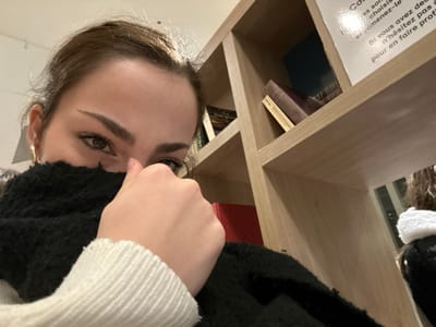 A young woman partially hides her face behind a black scarf and her hand. She has light brown hair pulled back, exposing gold hoop earrings. Her eyes are green, and her eyebrows are well-defined. Behind her is a light wooden bookshelf with various books and a sign with French text. The sign reads "Comes sol choisi- samenez-le Si vous avez de n'hésitez pas pour en faire prof". The overall image has a warm, slightly upward angle.