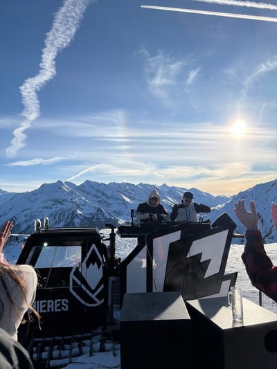 The image depicts an outdoor DJ event occurring at a high altitude in a snowy mountain environment within Schwendau, Austria. The scene is dominated by a clear blue sky, featuring a bright sun positioned in the upper right quadrant and two distinct white contrails; one straight and extending from the upper right, the other wavy and dissipating in the upper left. A majestic range of snow-covered mountains forms the backdrop.

At the center, a black tracked vehicle, likely a modified snow groomer, serves as a DJ stage. This vehicle bears the partial branding "IERES" and a white logo depicting a mountain peak above two horizontal lines, consistent with ski resort branding such as "HINTERTUX." Two male individuals, dressed in winter jackets, hoodies, and headwear (one with a white hoodie and cap, the other with a dark cap), are positioned behind a DJ console on a raised platform atop the vehicle, actively operating mixing equipment.

In the foreground, partially visible audience members are present. On the left, a person with long hair, wearing a light-colored hoodie, has a hand raised. On the right, a person in a red and black plaid shirt has both hands raised in an upward gesture. Black speaker units are situated on the snow-covered ground in front of the stage, and a clear glass beer stein rests on one of these speakers. The ground surrounding the setup is covered in snow. The scene is brightly illuminated by direct sunlight, casting distinct shadows.