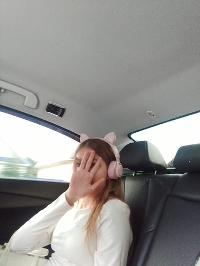 The image shows a young Caucasian female sitting in the back seat of a vehicle, presumably in the city of Pugachyov, Russia. She is wearing pink headphones with cat-ear attachments and a white long-sleeved shirt. Her right hand is held up, covering the right side of her face. The interior of the car, including the headrests of the back seats and the ceiling, is visible. Sunlight streams in from the windows, creating bright areas and shadows. The focus is on the person, with the background slightly blurred.