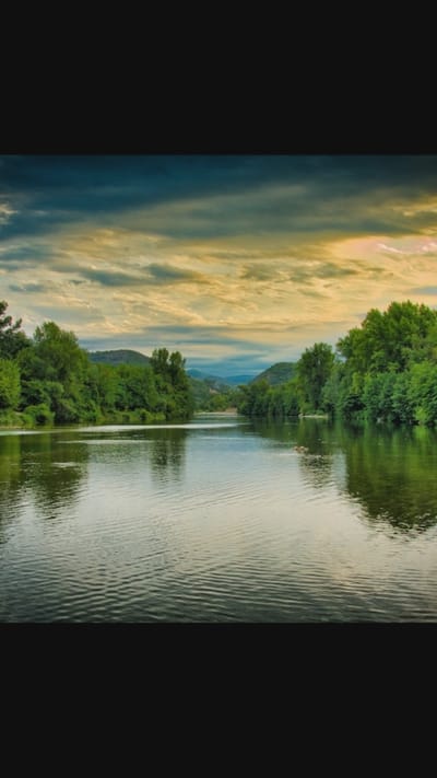 The image depicts a serene natural landscape, likely a river flowing through a verdant, tree-lined valley. The sky above is dramatic, with a mix of dark clouds and patches of golden light, suggesting either sunrise or sunset. The water's surface is calm, reflecting the surrounding trees and the sky. The lush green foliage on either side of the river indicates a healthy, well-watered environment. In the distance, rolling hills are visible, covered in trees, creating a sense of depth and vastness. There are no people or man-made structures visible in the scene, emphasizing the untouched, natural beauty of the location. The overall impression is one of tranquility and peacefulness.