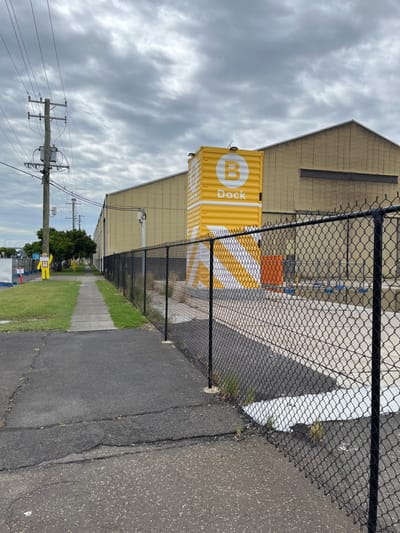 The image depicts an industrial or commercial area under an overcast sky, consistent with a location like Hamilton, Australia. A large, beige industrial building occupies the left background, featuring a distinct roofline and some darker horizontal elements on its facade.

A bright yellow shipping container stands vertically in front of the building, slightly to the right of the center. On its visible side, there's a white circle containing a bold capital 'B', and the word "Dock" written vertically below it in white. The container also has diagonal white and yellow stripes extending from it.

A black chain-link fence stretches horizontally across the middle ground, separating the industrial structures from an asphalt path or road in the foreground on the right. The asphalt shows visible cracks. A strip of green grass runs along the inner side of the fence.

In the lower left, a wooden utility pole with several cross-arms and numerous overhead power lines extends upwards into the frame. Further in the distance, to the left of the utility pole, several green trees and faint outlines of other buildings are visible. A small, yellow, human-shaped sign on a pole is vaguely discernible along the distant road.

The sky is entirely covered with thick, grey clouds, indicating an overcast day with diffused lighting and no harsh shadows. The overall scene suggests a functional, non-residential environment, possibly a port or a warehousing district.