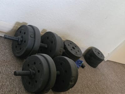 An indoor scene in Shotton, United Kingdom, depicts exercise equipment positioned on a speckled brown carpet against a textured white wall. The wall exhibits discoloration and staining along the white skirting board. In the foreground, a black adjustable dumbbell is partially visible, featuring a threaded bar and black weight plates secured by a spinlock collar. To its right, another black adjustable dumbbell, similarly constructed with threaded bar, spinlock collars, and black weight plates (some faintly marked "2.5KG"), rests on the carpet. Further right and against the wall, a stack of additional black weight plates with circular cutouts is present. A small blue and green microfiber cloth and a dark cylindrical object lie on the carpet between the assembled dumbbell and the stack of plates. The overall lighting is even, suggesting ambient indoor conditions. No individuals are present.