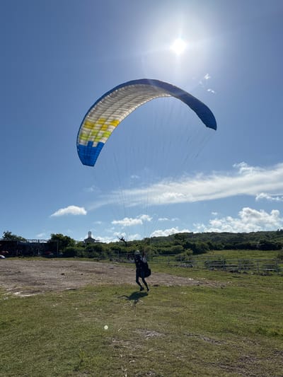 A wide-angle shot captures a paraglider taking off in Benoa, Indonesia. The blue and yellow canopy is prominently featured against a bright blue sky with scattered clouds and the sun positioned at the top-left. A person is on the ground, appearing to be running to gain momentum for the launch. The foreground consists of a grassy area transitioning to a dirt path. In the background, trees and a fence line are visible.