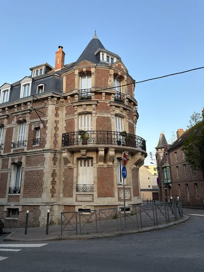 The image depicts a street corner in Dieppe, France, characterized by a prominent, multi-story brick building with a conical roof and decorative balconies. The architecture suggests an early 20th-century style. The building features red brickwork interspersed with light-colored stone accents and white-shuttered windows. A chimney rises from the roof. In the background, other buildings of similar brick construction are visible, along with a street with a pedestrian crossing and metal safety barriers. Traffic signs, including a "no entry" sign and a directional arrow sign, are present on a pole near the corner. The sky is clear and blue. There are no people or vehicles actively engaged in actions within the frame. The scene is quiet and appears to be captured during daylight hours, likely late afternoon or early morning due to the soft lighting.