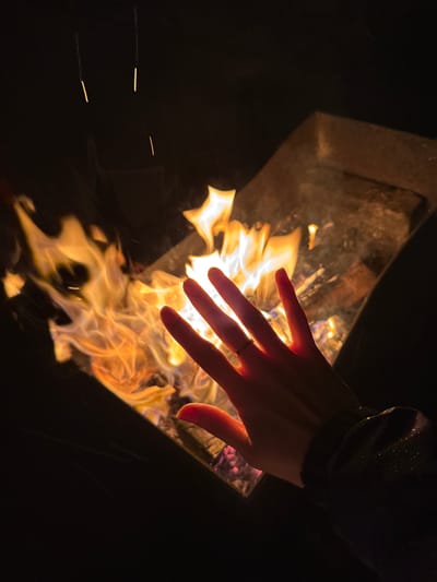 A hand with a ring on its ring finger is extended over a fire burning in a metal container. The fire is the primary light source, illuminating the hand and the immediate surroundings. A dark-colored sleeve is visible in the upper right. The environment is mostly dark, suggesting an outdoor setting at night. The image does not contain sufficient information to verify the location in Mayak, Russia.