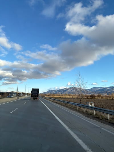 The image depicts a multi-lane highway in Akhalsopeli, Georgia, under a partially cloudy blue sky. The perspective is from a vehicle moving forward in the rightmost lane. Directly ahead, a dark-colored semi-truck tractor unit, without a trailer, is proceeding in the same lane. To the left, an elevated section of the highway features multiple lanes with several vehicles, including large white buses and cars, moving in the opposite direction. Streetlights with curved arms line the left side of the road. To the right of the road, a metal guardrail separates the highway from a wide, tilled agricultural field of brown soil. A single, leafless deciduous tree stands prominently within this field, beyond the guardrail. In the distant background, snow-capped mountains extend across the horizon beneath scattered white and grey clouds. No individuals are explicitly visible or performing actions outside of operating vehicles.