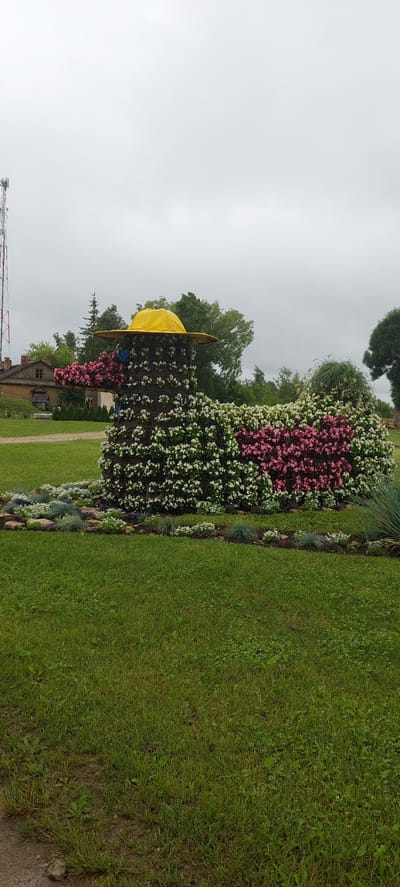 The image shows a flower sculpture of a duck in a park in Linkuva, Lithuania. The duck sculpture is made of wire mesh and covered with pink and white flowers, with a yellow hat on its head. The duck is standing on a bed of green grass, with a building and trees in the background. The sky is overcast.