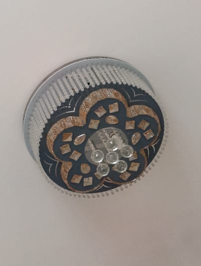 A ceiling-mounted light fixture is centered against a plain white ceiling in Xojeli, Uzbekistan. The fixture is circular, featuring an outer silver-toned ring adorned with vertical, translucent white rectangular elements. The central decorative element is a dark blue, four-lobed shape, bordered by gold-toned glitter and inlaid with numerous clear, faceted, square crystals. A cluster of clear, spherical, faceted crystals is suspended from its core, functioning as the primary light diffuser. No individuals or additional objects are visible within the frame.
