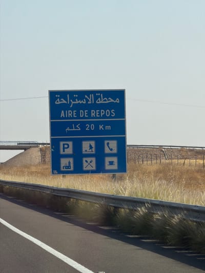 A roadside photograph, likely taken from a vehicle, captures a blue sign indicating an "Aire de Repos" (Rest Area) 20 kilometers ahead. The sign features both Arabic and French text, accompanied by pictograms illustrating services available at the rest area: telephone, fuel, picnic area, restaurant, parking, and possible lodging. The sign is situated alongside a highway, with roadside vegetation and a metal guardrail visible. The location is suggested to be near Nzalat Laadam, Morocco.