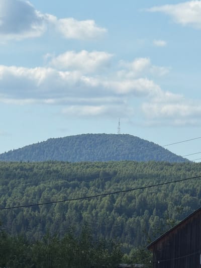 The image depicts a panoramic view of a forested mountain range under a partly cloudy sky. In the foreground, the edge of a rustic wooden building is visible on the right side. The middle ground is dominated by a dense, green forest that covers rolling hills. The background features a larger, rounded mountain also covered in trees, with a slender transmission tower visible near its summit. The sky is a pale blue, with scattered white clouds. There are no people or animals visible in the image. The scene appears to be a natural landscape, possibly in a rural or mountainous area. This image does not contain any specific landmarks or details that would allow for identification of its location within Krasnovishersk, Russia.