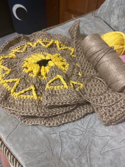 The image shows a close-up view of a crochet project, likely a sunflower motif, made with brown and yellow yarn. The project is laid out on a textured surface that appears to be a tablecloth or some other fabric with a floral pattern. To the right of the crochet work is a spool of brown twine and a ball of bright yellow yarn. In the background, a dark blue object with a white crescent moon symbol is visible. The overall scene suggests a crafting or hobby activity. There is no indication of the city of Zmiiv, Ukraine, or any specific urban environment.