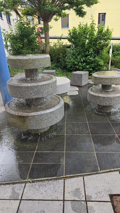 The image depicts an outdoor decorative fountain in Zirndorf, Germany, under bright daylight. The fountain is composed of a series of granite-like stone elements arranged linearly across a tiled ground. Several large, circular, flat stone discs, resembling millstones, form part of the fountain, with water continuously flowing over their surfaces and cascading downwards. Interspersed among these circular elements are smaller, square or rectangular stone blocks.

The water collects and flows through dark grey tiled channels on the ground, creating ripples and reflections. Adjacent to the fountain's tiled channels is a lighter grey tiled pathway, showing some areas of dampness.

In the background, a prominent light yellow building with multiple windows is visible. Some windows feature distinct blue frames. Lush green foliage, including several trees and dense bushes, grows between the fountain and the building, adding a natural element to the scene. A robust tree trunk with branches extends from the lower left towards the upper background. The overall impression is one of a peaceful, well-maintained urban or park setting. There are no people or discernible text visible in the image.