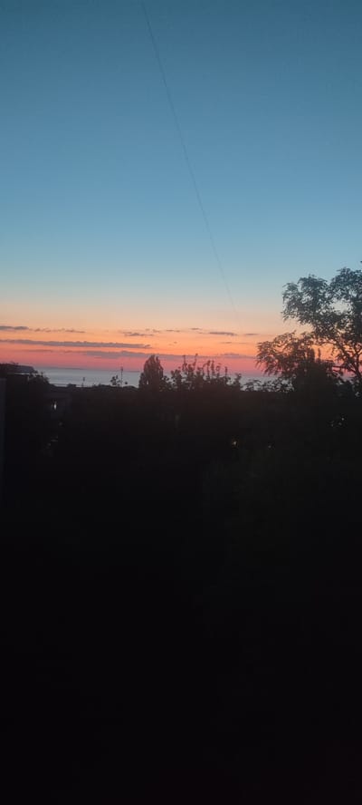 The media file depicts a natural scene at dusk, with the sky transitioning from a clear blue at the top to a gradient of orange and pink hues near the horizon, indicating sunset or sunrise. A thin, dark line, possibly a wire or a contrail, stretches diagonally across the upper portion of the sky. Below the colorful sky, a body of water, likely a river or sea, is visible, with a faint silhouette of land and structures in the distance, suggesting a coastline or the opposite bank. The foreground is dominated by dark silhouettes of trees and buildings, obscuring most details of the lower landscape. Some faint lights are visible within the darkened structures, hinting at human habitation. The overall mood is serene and tranquil, evoking the quiet beauty of twilight. No text is visible in the image. The location context of Svitlovodsk, Ukraine, suggests this may be a view of the Dnieper River or the Kremenchuk Reservoir.
